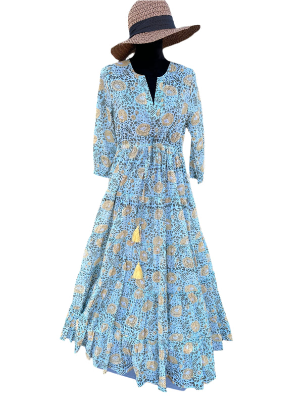 The dress in the image has a light blue base color with beige, white, and black floral patterns. There are also small yellow tassel accents. The overall color palette is soft, earthy, and vintage-inspired, giving it a timeless, bohemian look